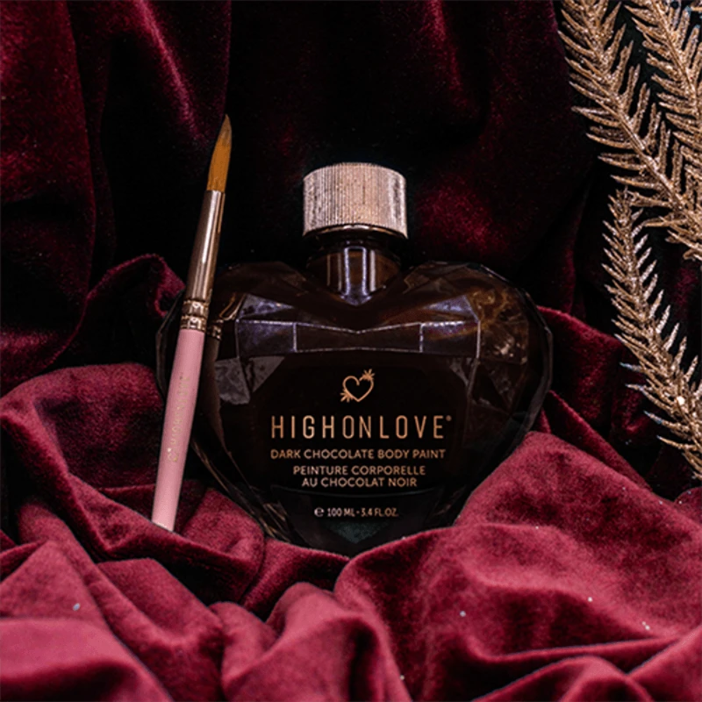 A bottle of Dark Chocolate Body Paint by High On Love sitting next to it's paint brush on a red cloth.