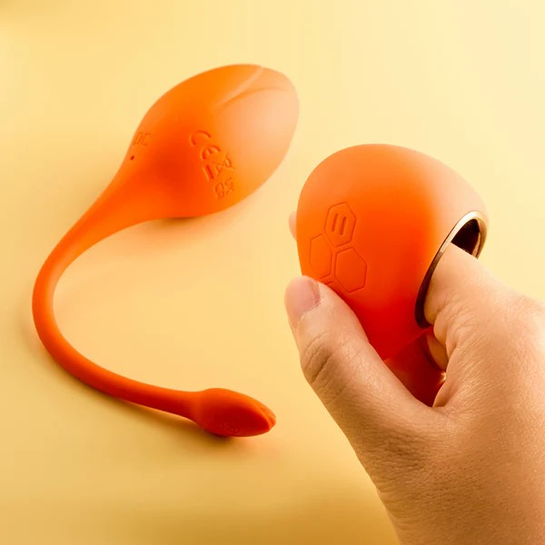 A orange Lili vibrator against an orange background and a hand holding the remote.