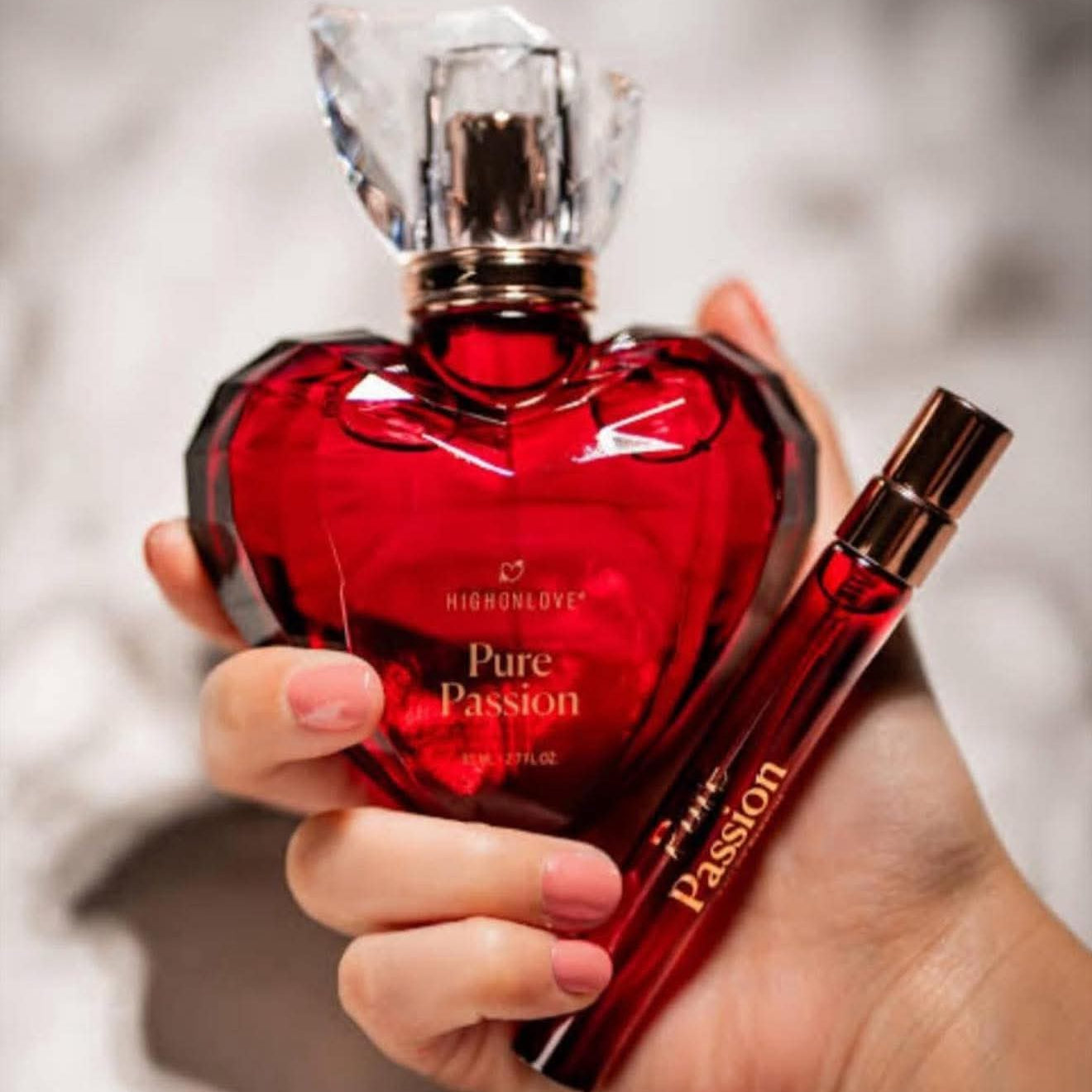 Red heart-shaped perfume bottle and travel sized bottle labeled 'Pure Passion by High On Love' held by a hand against a blurred background.