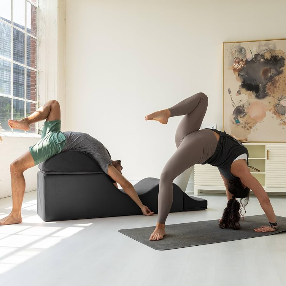Two people practicing yoga with the black aria chaise in a bright room with large windows.