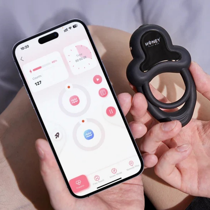 A black Anello Honey Play Box cock ring being held next to a phone showing the app that controls it.