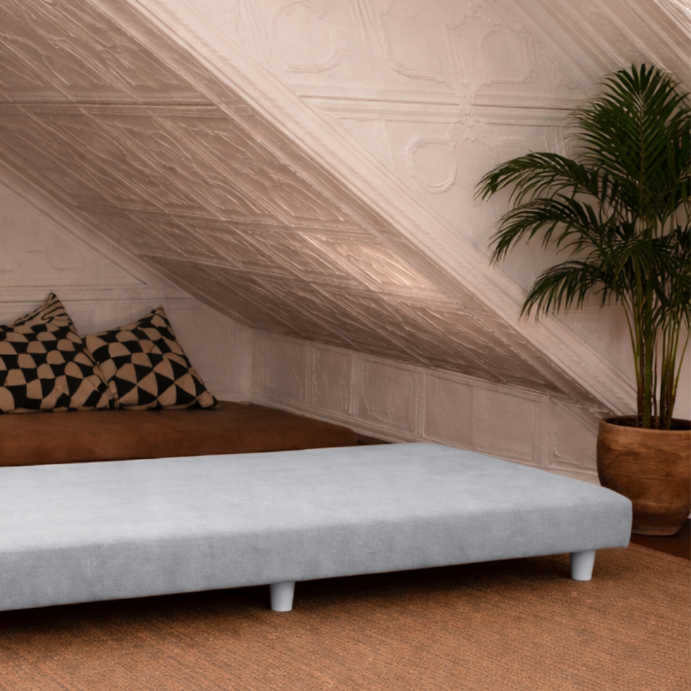 Unfolded divan daybed under a sloped ceiling with a plant and cushions in a room.