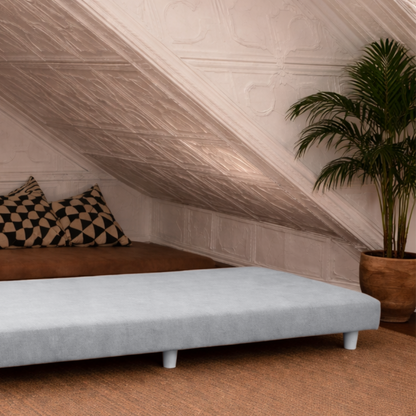 Unfolded divan daybed under a sloped ceiling with a plant and cushions in a room.