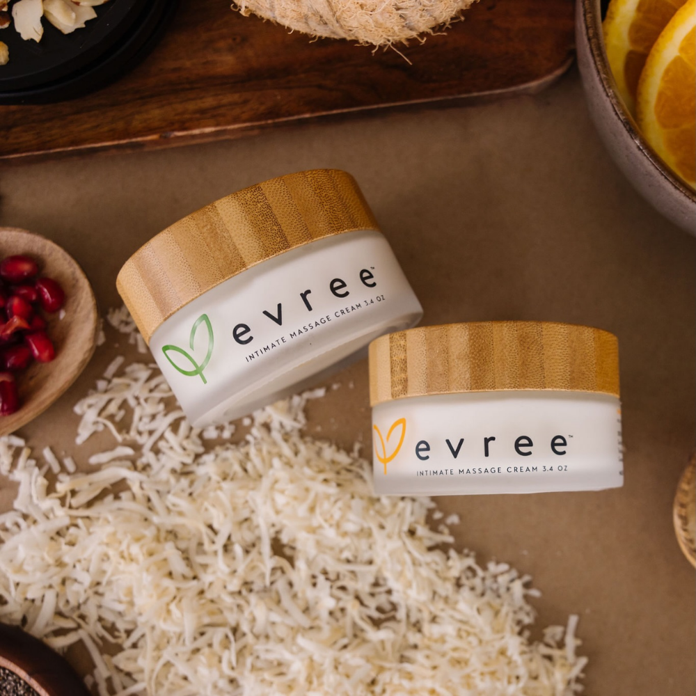 Two white jars of Edible Massage Cream by Evree, one with a green leaf on it and one with a yellow leaf on it, sitting on a brown table.