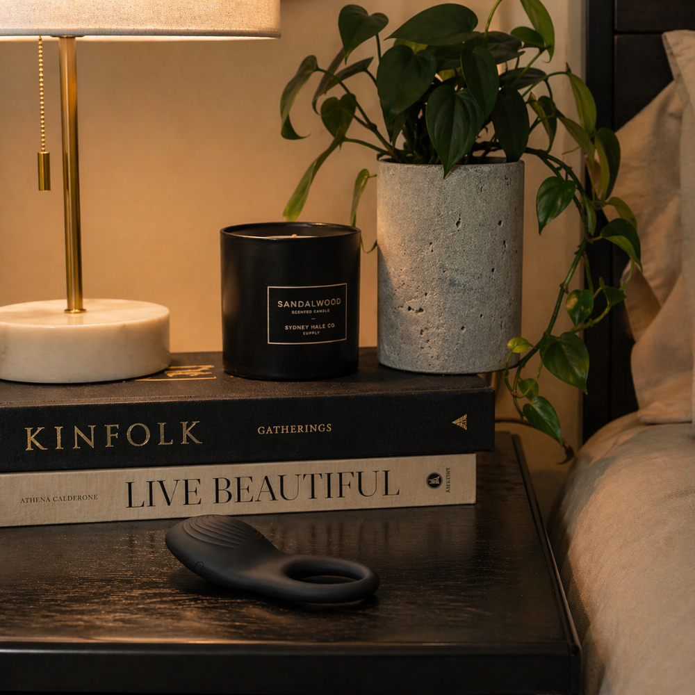 A black Imprint Cock Ring by Envy sitting on a bedside table next to a light and plant.