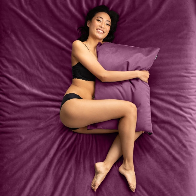 A lady laying on an aubergine bed hugging a Aubergine Fascinator pillow, Bedroom Furniture by Liberator.