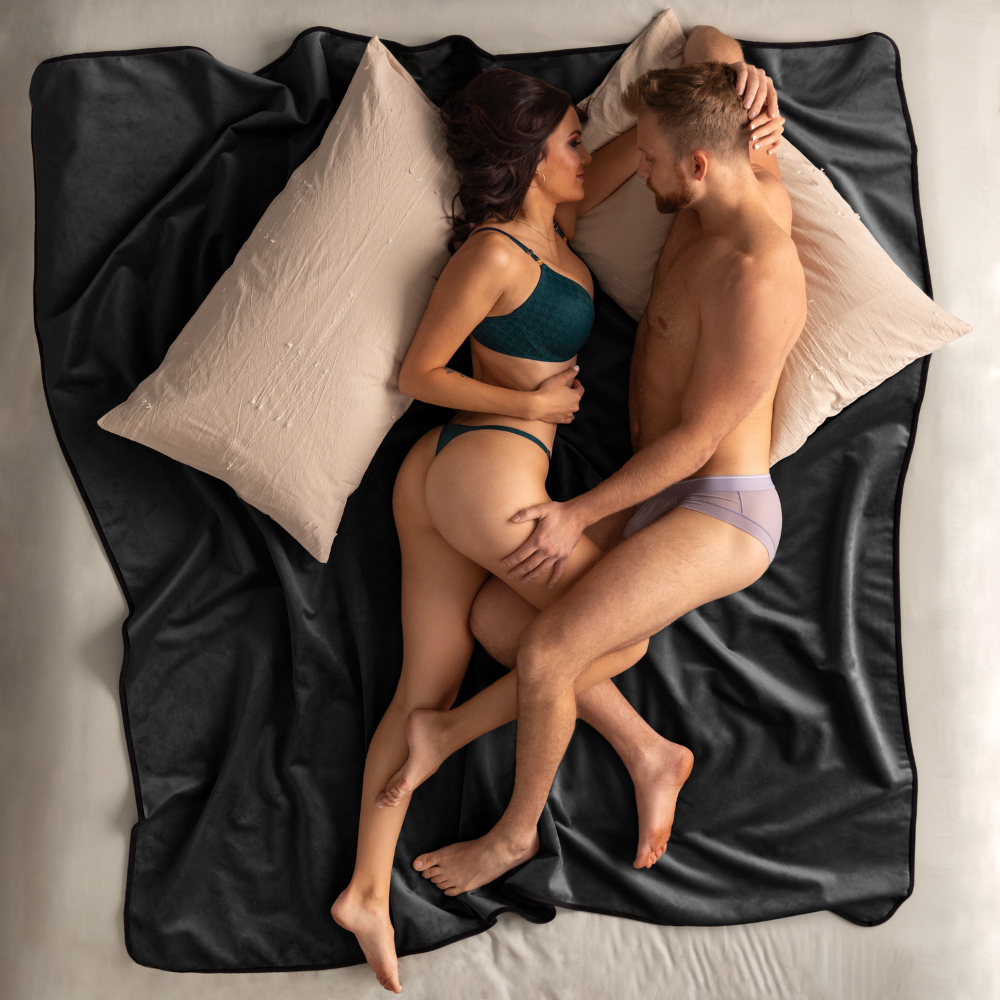 A couple in underwear on a King size black Fascinator Splashproof Throws, Bedroom Furniture by Liberator sitting on a bed.
