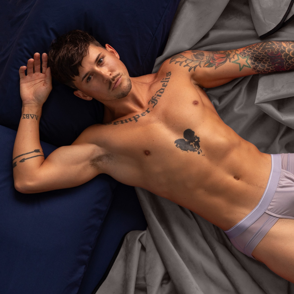 A man in underwear laying on a grey Fascinator Splashproof Throws, Bedroom Furniture by Liberator sitting on a bed.