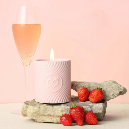 A pink massage candle mini by High On Love sitting next to a glass of champagne and strawberries.