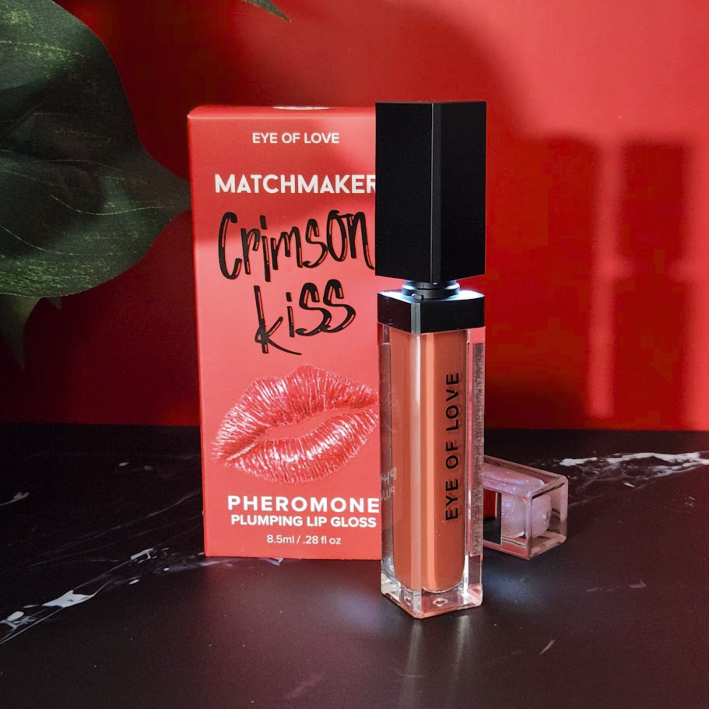 A red tube of plumping Crimson Kiss lip gloss by Eye of Love sitting next to the box against a red background.