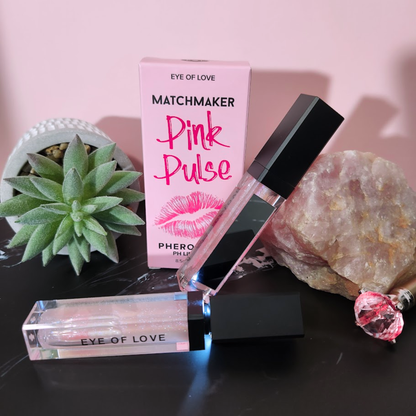 A two black and pink tubes of pheromone pH balanced Pink Pulse lip gloss by Eye of Love sitting next to the box, a crystal rock and a plant.