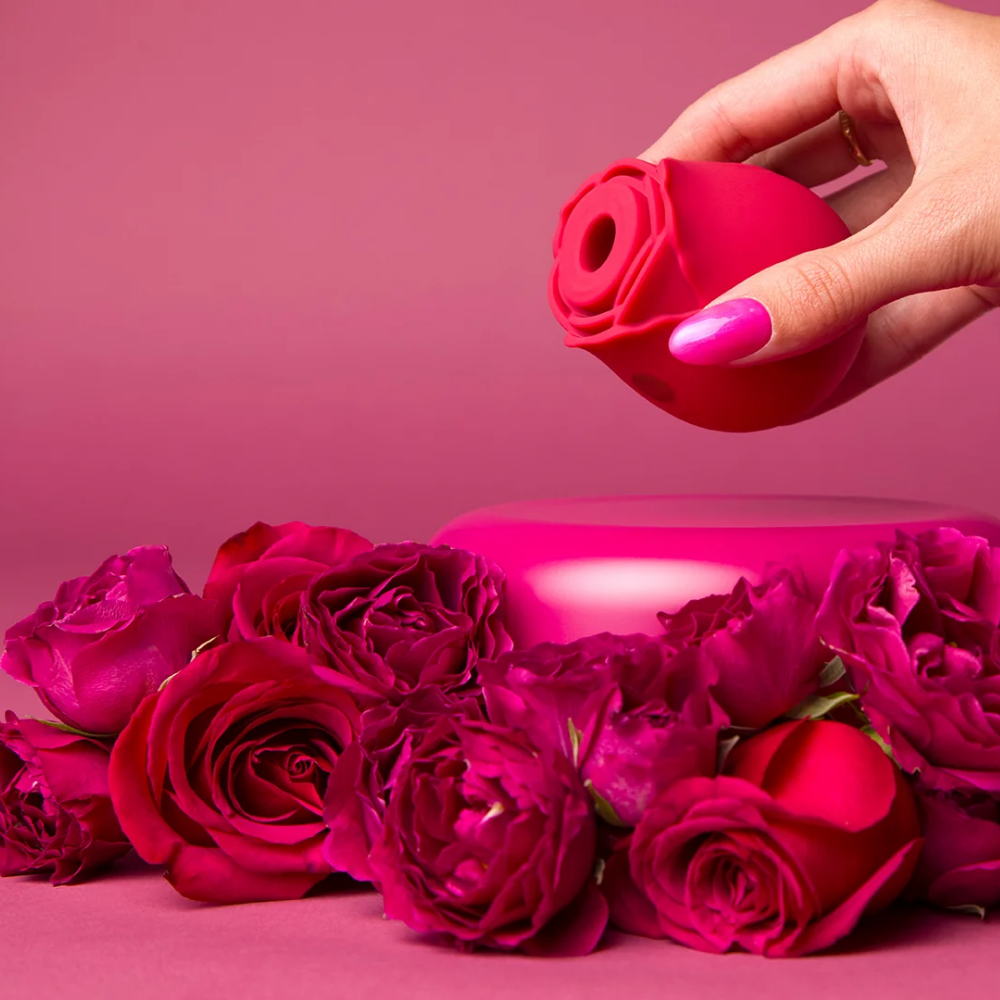 A hand holding a red Rosegasm Air Rose Clitoral Toy above a bed of red flowers.