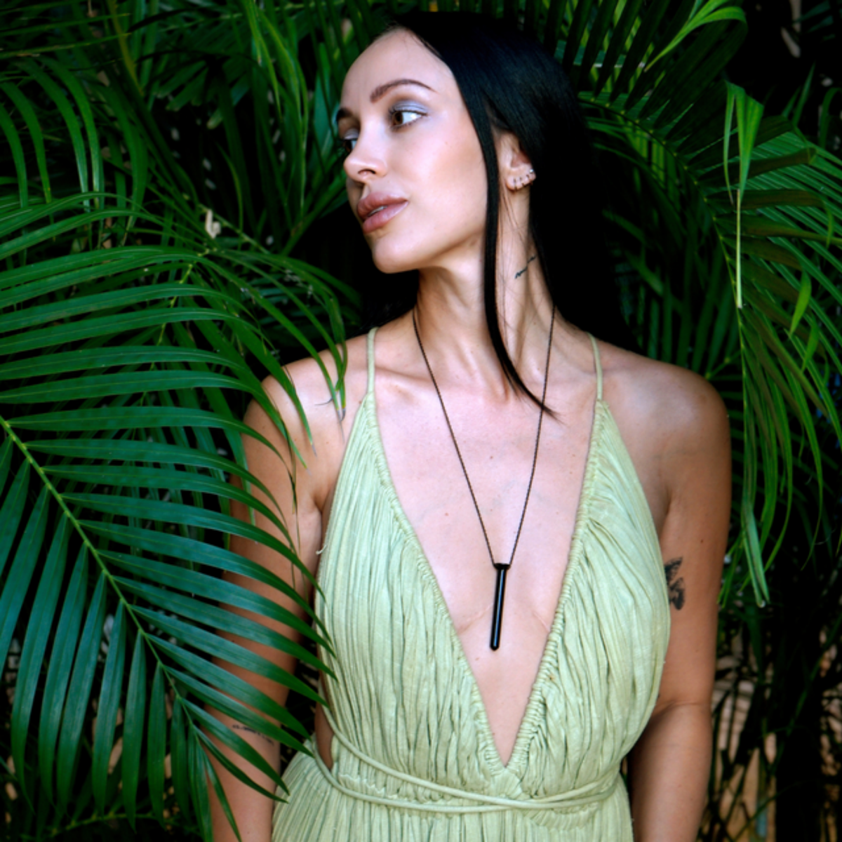 A lady wearing a black Vesper mini necklace surrounded by green leaves.