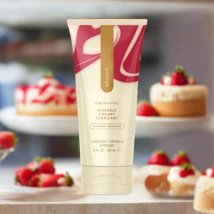 A cream colored tube of Strawberry Cheesecake Whipped Lubricant by Euforia against a background of strawberry cheesecakes.